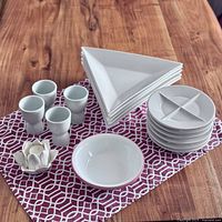 White dish lot including 5 triangle plates, 6 fondue divided plates, 4 egg cups, 2 bowls, and 1 lotus candle holder displayed on a patterned cloth on wooden surface.