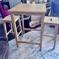 Photo of light wood square bistro table with two matching stools showing overall set, wear visible on stool seats.