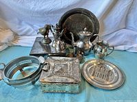 Photo showing all mixed metal decor items including tea set, trays, box, candle holder, bird figurine and basket arranged on a blue surface with white backdrop.