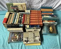 Overview of approximately 45 hardcover books arranged in rows on a blue surface, showing various titles, color bindings, and conditions.