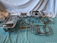 Wide view of six different metal casserole cradles and racks arranged against a cloth background. Includes cradle with removable cover, wire racks with wooden handles, and various round and rectangular shapes.