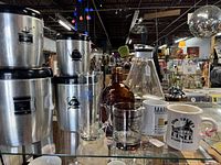 Photo of metal West Bend canister set with black lids, a textured glass tumbler, brown vintage glass bottle, two ceramic mugs, and a glass carafe with metal holder.