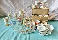 Various vintage glassware including mugs, souvenir mugs, shot glasses, pitchers, and glass cups arranged on table.