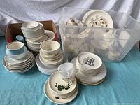 Overview of varied vintage dishware including plates, bowls, cups stacked and contained in clear plastic bin and cardboard box