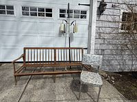 Front view of wooden sofa frame, kitchen chair, and tension pole lamp placed outside on concrete with garage door background.