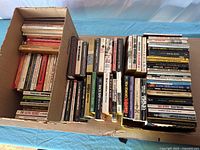 Wide shot of approximately seventy paperback books arranged in open cardboard box and stacked beside it showing various spine titles and conditions.