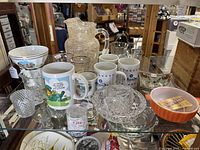 Wide shot showing a variety of glassware, ceramic mugs, bowls, and crystal items on a glass shelf.