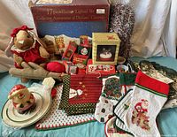 Full lot overview showing textiles, Dickens village set box, plush doll, mugs, stockings, and candle, all arranged on a bedspread.