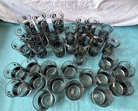 Grouped view of over 35 glasses in varying heights with black bands and gold coin designs on clear glass.