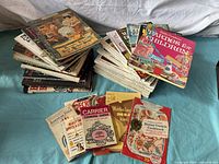 Wide view of two stacks of vintage cookbooks and cooking cards showing variety in titles and conditions.