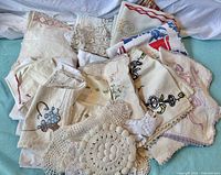 Large assortment of vintage linens piled together showing varied embroidery, crochet doilies, and different textures and colors, including white, cream, and pastel tones.