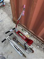 Three children's metal scooters shown lying on the ground next to a shipping container, each in used condition.