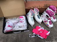 Three pairs of children's skates and pink protective gear laid out, showing different types: figure skates, inline skates, and strap-on skates.