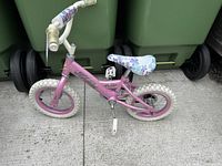 Pink Nakamura girls bicycle with white 12.5" tires, floral seat and white handlebars leaning against green bins.