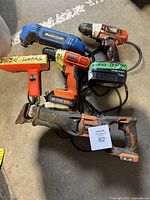 Five assorted power tools photographed together on concrete floor. Shows wear and dirt consistent with construction use. Includes Mastercraft rotary tool, Ryobi cordless drill, Ridgid reciprocating saw, and others.