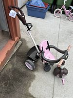Side view of the pink and brown Little Tikes push trike leaning against a container. Shows the seat, three wheels, pedals, and the push handle.