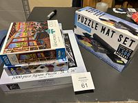 Photo showing three boxed jigsaw puzzles and boxed puzzle mat set on table.