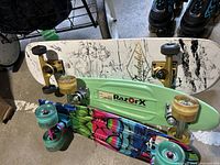 Top image showing three skateboards stacked. White deck skateboard with black artistic graphics and signature is on top, featuring yellow trucks and aged wheels. Below are two plastic cruiser skateboards with bright colors, one mint green with blue trucks and multi-colored wheels, the other lime green with matching wheels and trucks.