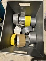 Top-down photo of plastic bin containing multiple partially used rolls of duct tape, masking tape, and other adhesive tapes in various colors including silver, yellow, gray, white, and beige.