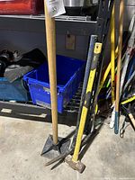 Photo showing the tamper plate with wooden handle, the yellow-handled sledge hammer, and the black-handled pick axe standing on a concrete floor near shelving.