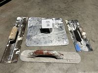 Lot of five construction trowels: two large wooden-handled trowels, one smaller wooden-handled trowel, one metal handled trowel with paint, and a large square aluminum hawk with label visible.