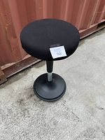 Full view of the black wobble stool showing round cushioned seat, central black and silver column, and round black weighted base on concrete ground near a container.