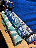 Four rolled woven beach mats in teal, blue, cream, and purple with floral and striped patterns, secured by straps, displayed on a wooden surface with a blue background.