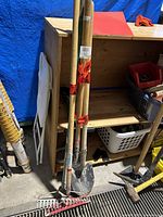 Wooden handled yard tools including two shovels and two red rakes leaning against wood shelving unit, showing wear and usage dirt