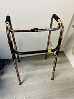Front angled view of bronze metal walking frame with black grips on handles, adjustable legs, and rubber tips.