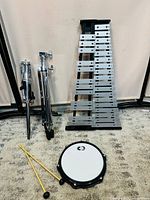 Photo showing glockenspiel laying on black frame with metal bars, two chrome stands, a small drum with CB logo and two yellow drumsticks on carpeted floor.