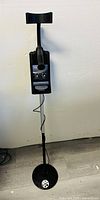 Full view of metal detector standing against white wall, showing armrest, control box, and search coil.