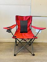 Front view of red folding camping chair showing seat, armrests with cup holder on right side, and metal frame.