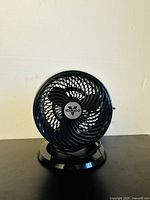Front view of small black Vornado desk fan on round base, visible cage and blades, Vornado logo center.
