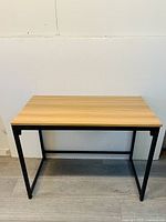 Front view of a rectangular office side table with a light oak wood top and matte black metal legs against a white wall on a grey floor.