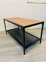 Three-quarter view showing wooden tabletop, black metal legs and mesh shelf underneath.