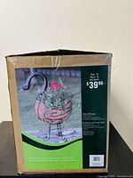 Photo of cardboard box packaging showing an image of the metal goose-shaped planter with flowers placed inside it outdoors, with price tag visible.