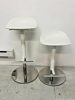 Front and side view of two white Janinge bar stools showing adjustable height levers, curved seats, and circular chrome bases with footrests.