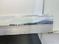 Long rectangular wall art photographed standing on black floor against white wall, shows ocean scene with ships and distant mountains.