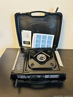 Open portable gas stove inside its carry case with instruction manuals placed inside the case lid.