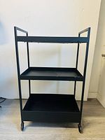 Front view of black metal rolling kitchen cart showing three shelves and four wheels.