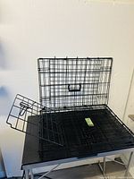Folded black wire mesh dog cage with door and carrying handle on top, laid flat on a table.