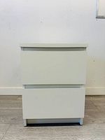 Front view of a white 2-drawer nightstand cabinet against a white wall, showing smooth flat drawer fronts without handles, and short integrated legs.