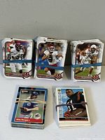 Three stacks of trading cards held with rubber bands, featuring football and baseball cards on a white table against a gray wall.