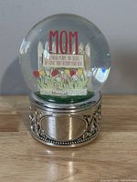 Front view of the snow globe featuring the MOM text and floral message inside, placed on an ornate metal base.