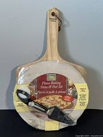 Photo showing round pizza stone wrapped in plastic with wooden peel behind it, handle labeled Talyan Villa