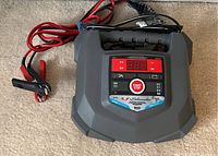Front view of Schumacher Electric SC1223 battery charger showing digital display, indicators, and attached battery clamps.