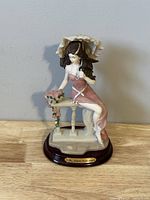 Front view of porcelain figurine of lady with umbrella seated on stone bench with flowered table, mounted on wooden base with nameplate