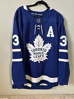 Front view of blue Toronto Maple Leafs hockey jersey with white maple leaf logo, "A" on chest, and sleeved white stripes.