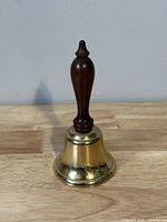 Side view of the vintage brass hand bell showing brass bell and wood handle standing upright on wooden surface.