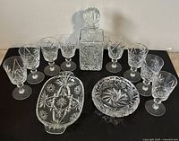 Photo showing 12 cut crystal vine glasses, a square decanter with stopper, an oval ashtray, and a round ashtray or dish all with intricate starburst and floral cut glass patterns.